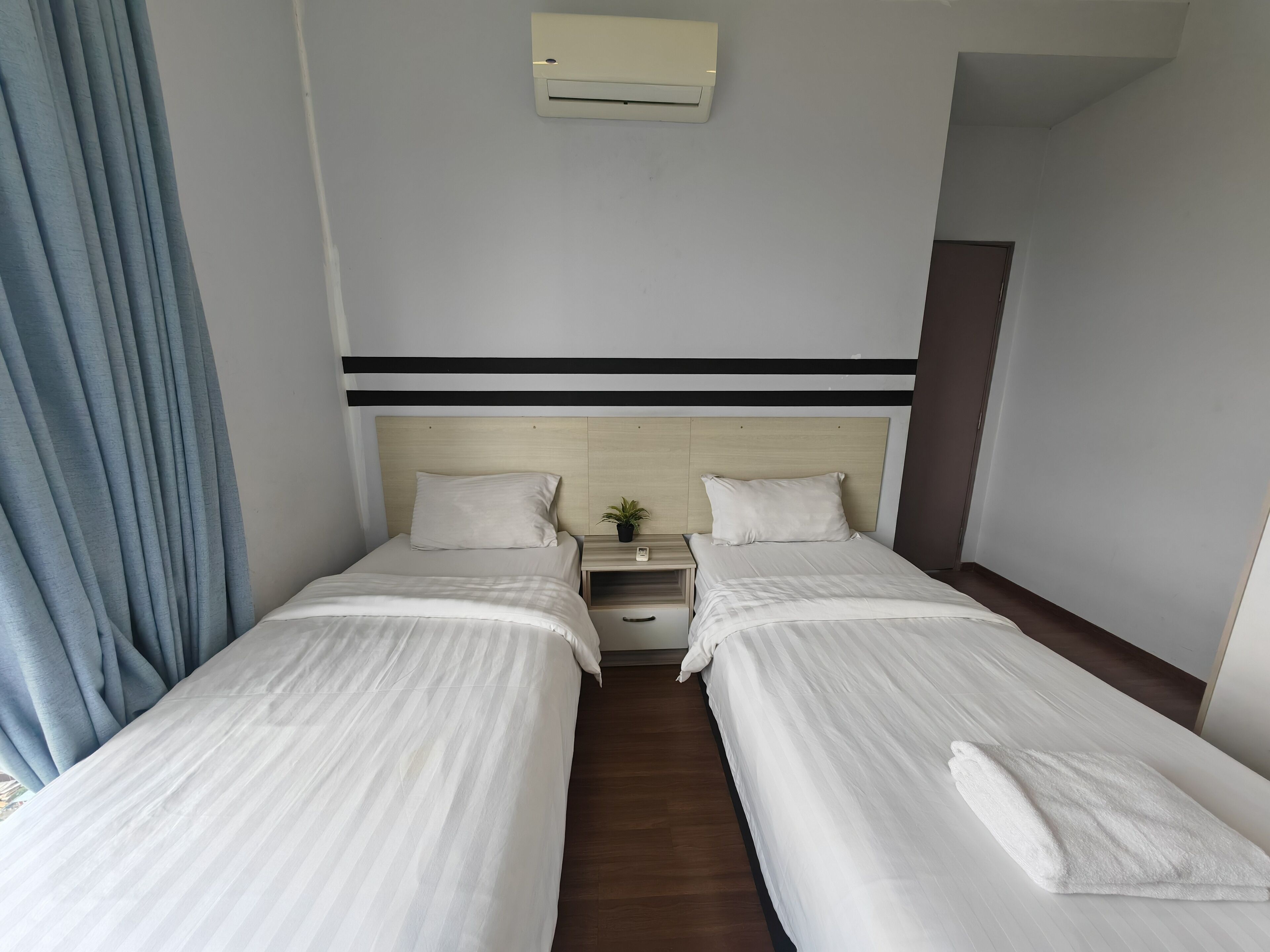A hotel room with two single beds, a bedside table with a lamp, a small potted plant, and a wall-mounted air conditioner.