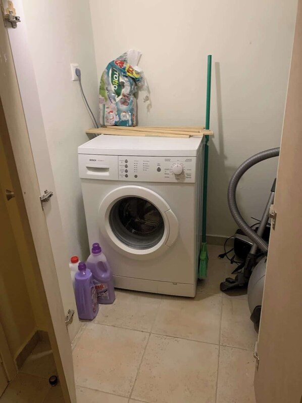 A compact laundry room with a front-loading washing machine, a vacuum cleaner, and cleaning supplies. - Cassiopeia 1BR Apt on Beach with Pool (Pyrgos Psilonerou)