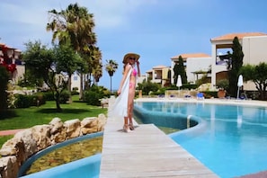Une piscine entourée d’un ciel dégagé, de palmiers et d’un bâtiment doté d’un toit en tuiles.
