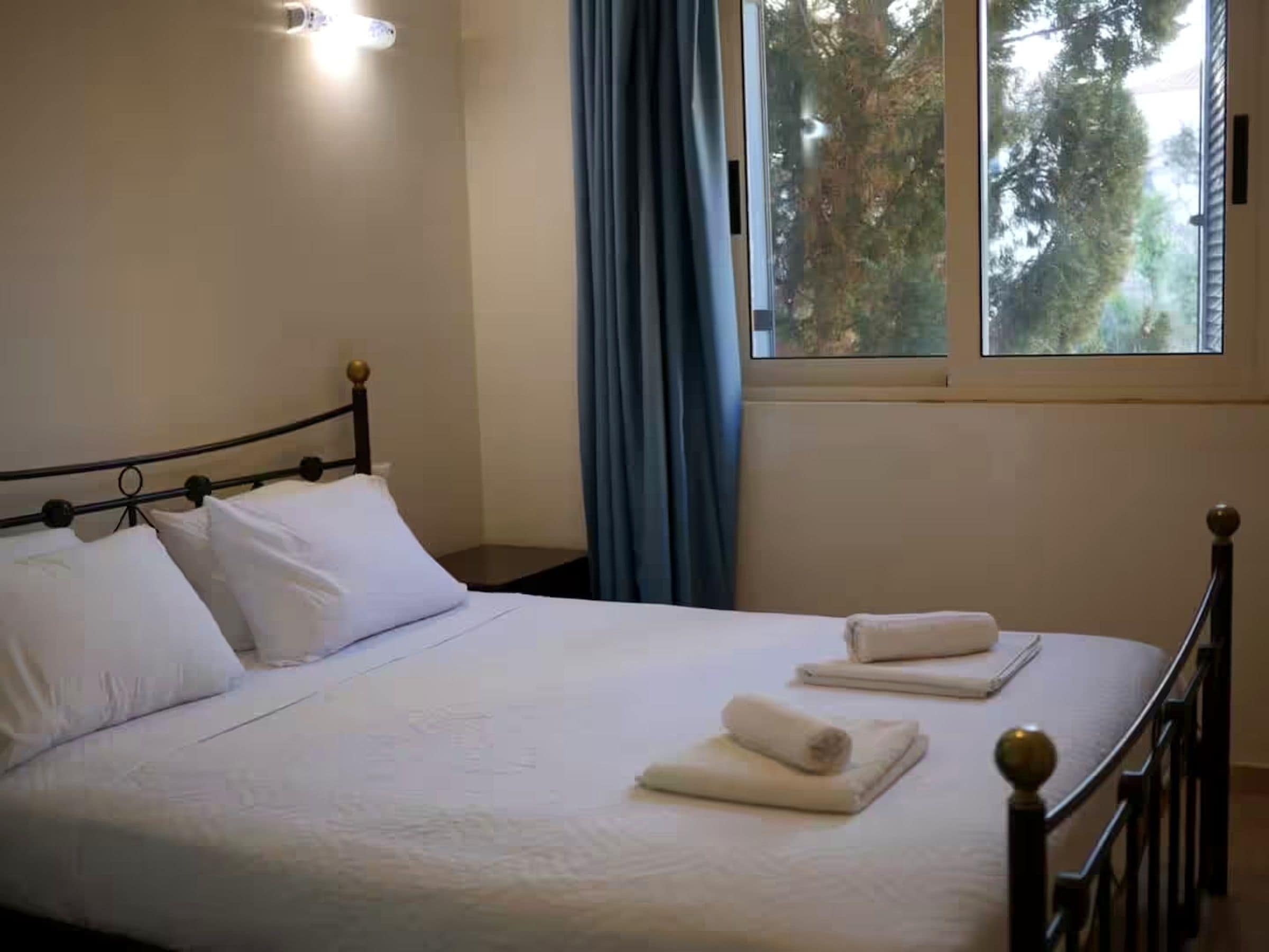 A neatly made bed with white linens and two towels, a window with blue curtains, and a wall-mounted light fixture.