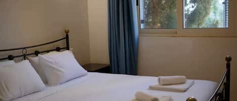 A neatly made bed with white linens and two towels, a window with blue curtains, and a wall-mounted light fixture.