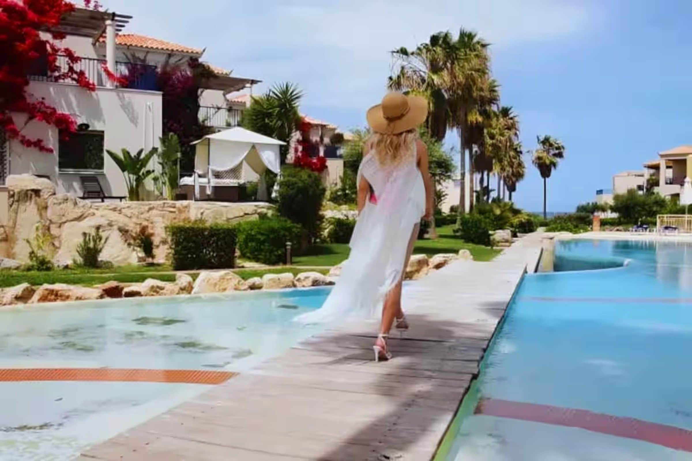 Une personne vêtue d’une robe blanche et portant un chapeau de paille se promène le long d’un chemin bordant une piscine.