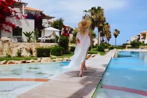 A person in a white dress and straw hat walking along a poolside path. - Cassiopeia 1BR Apt on Beach with Pool (Pyrgos Psilonerou)