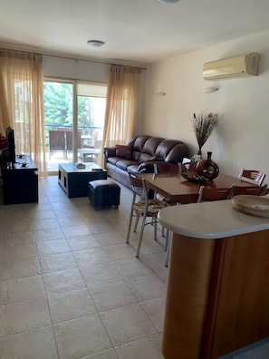 A living room with a sofa, dining table, chairs, and a view through a large window. - Cassiopeia 1BR Apt on Beach with Pool (Pyrgos Psilonerou)