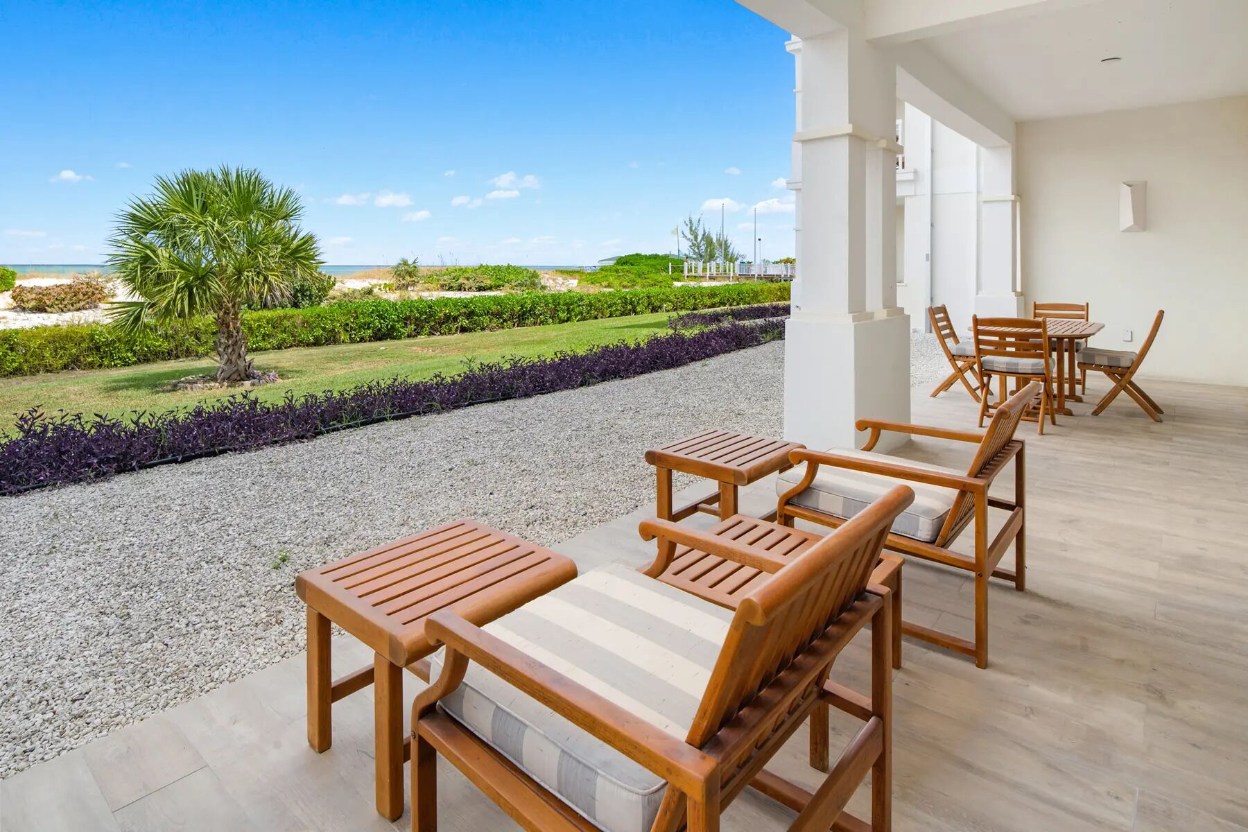 A patio with wooden furniture and a view of a landscaped garden and beach.