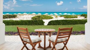 Uitzicht vanaf een balkon met twee houten stoelen en een kleine ronde tafel, op een strand met witte parasols en een helderblauwe lucht.
