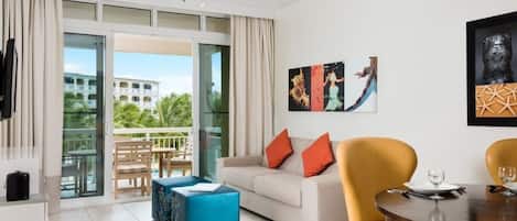 A modern hotel room with a sofa, a dining table, and a balcony with a view of a building and palm trees.