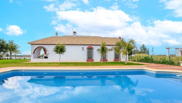 Un edificio blanco con techo de tejas, una piscina y un cielo azul despejado.