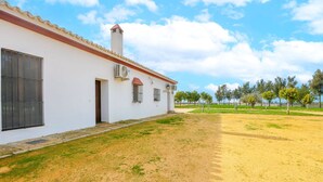 Un edificio blanco con techo rojo, una chimenea y una puerta pequeña. Hay un área verde al frente y una fila de árboles al fondo.