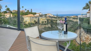 Un balcon avec une table et une chaise, donnant sur un paysage côtier avec des maisons et des palmiers.