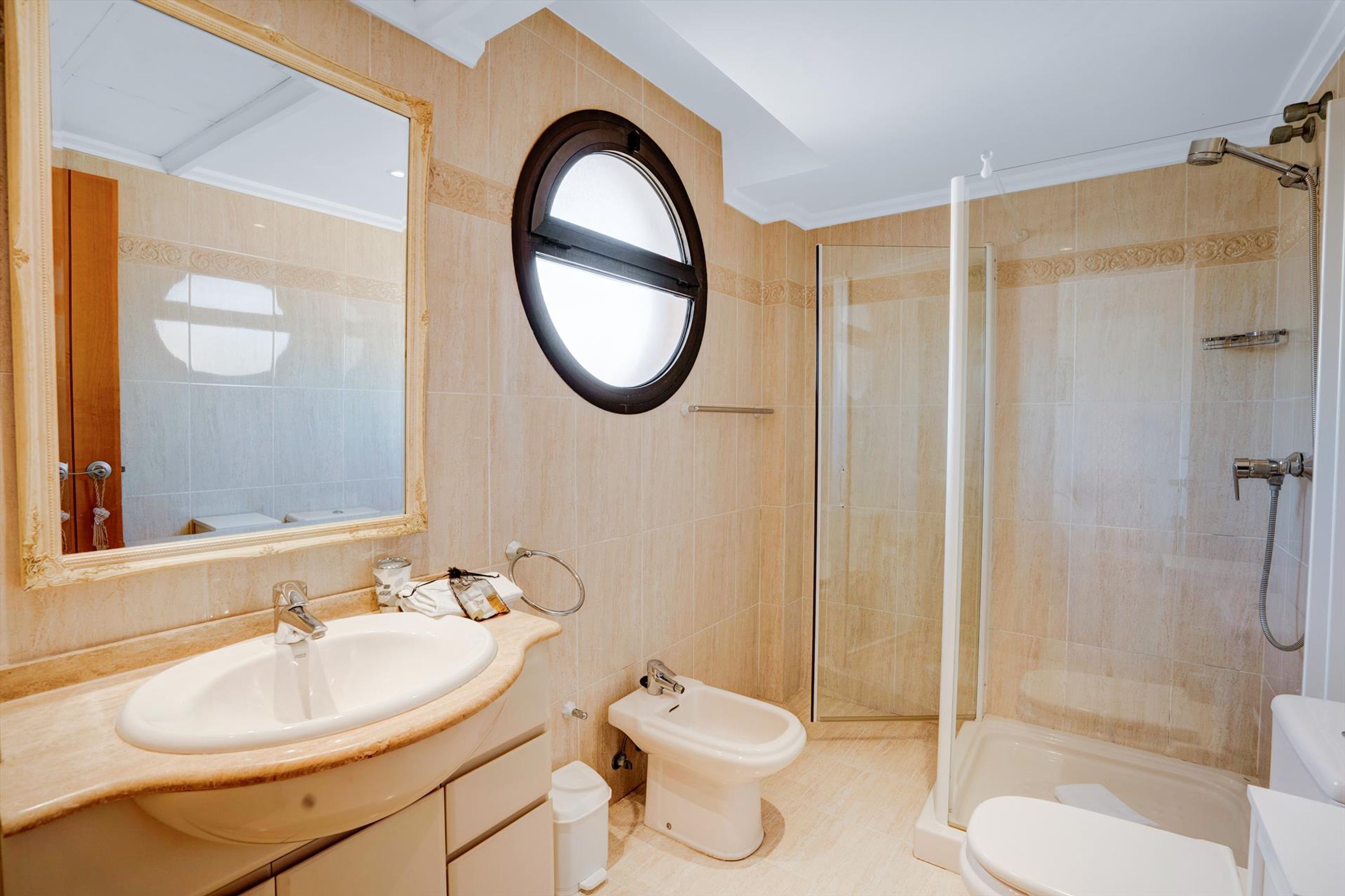 A bathroom with a large oval mirror, a sink, a bidet, a toilet, and a glass-enclosed shower.