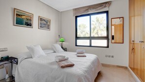 Un dormitorio con una cama grande, dos cuadros, una ventana con vistas, un armario y una mesita de noche con una lámpara.