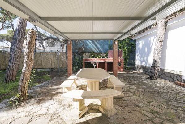 A covered patio with a stone table and benches, surrounded by trees and a brick wall.
