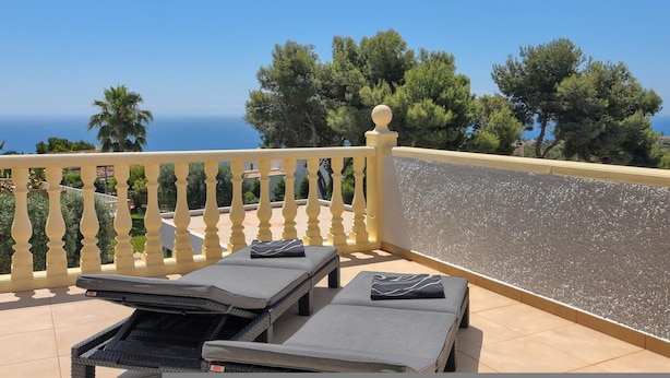 Ein Balkon mit zwei Liegestühlen, einer Brüstung und Blick auf das Meer und Bäume.