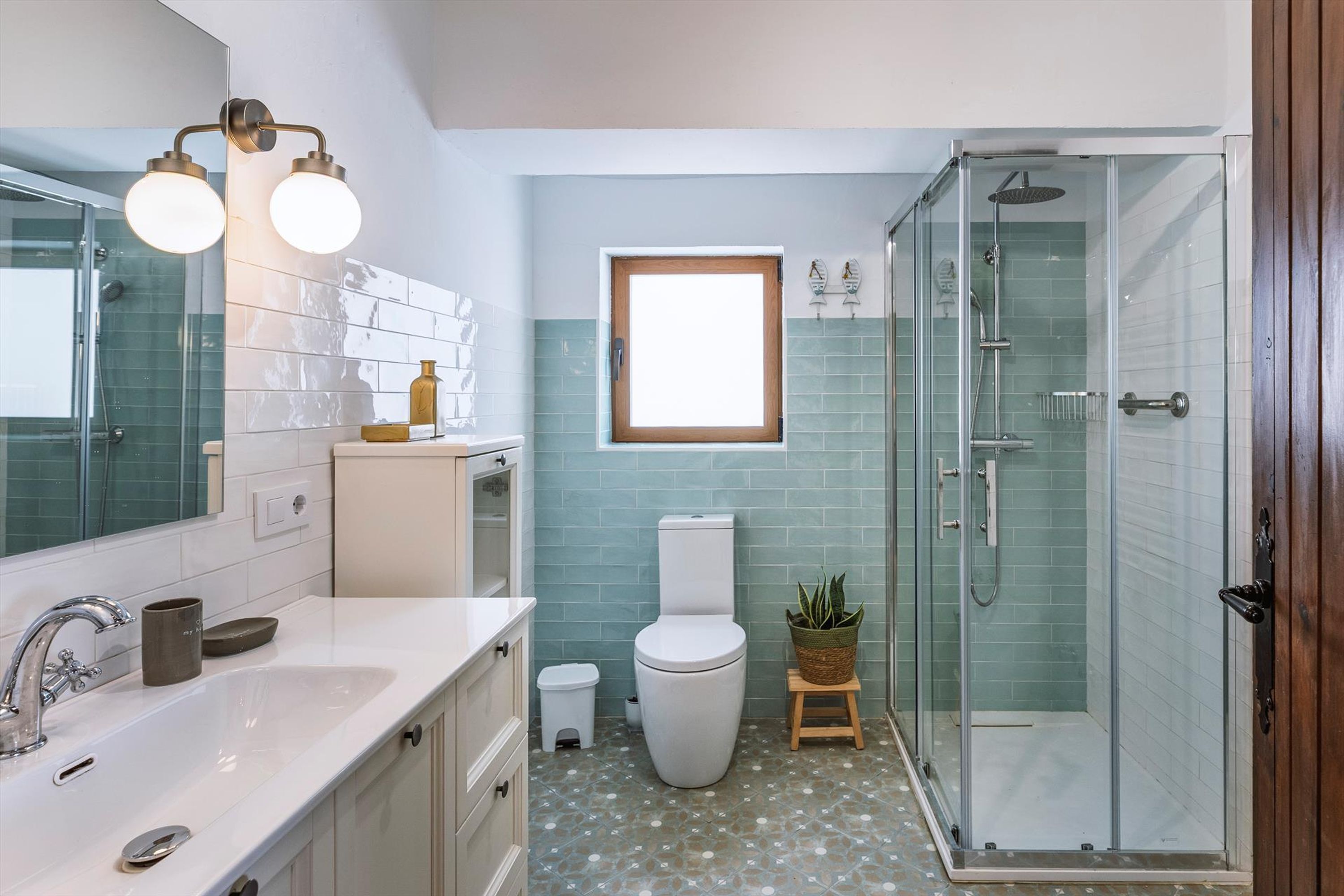 Une salle de bain moderne avec une douche à l’italienne en verre, un lavabo blanc avec un miroir et une petite fenêtre au-dessus des toilettes.