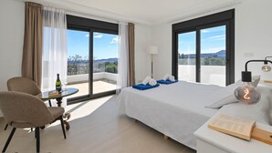 A modern hotel room with a large bed, a sitting area with a table and chair, and a view of the city through large windows. - Casa Renata 8 pax in Javea (Javea)