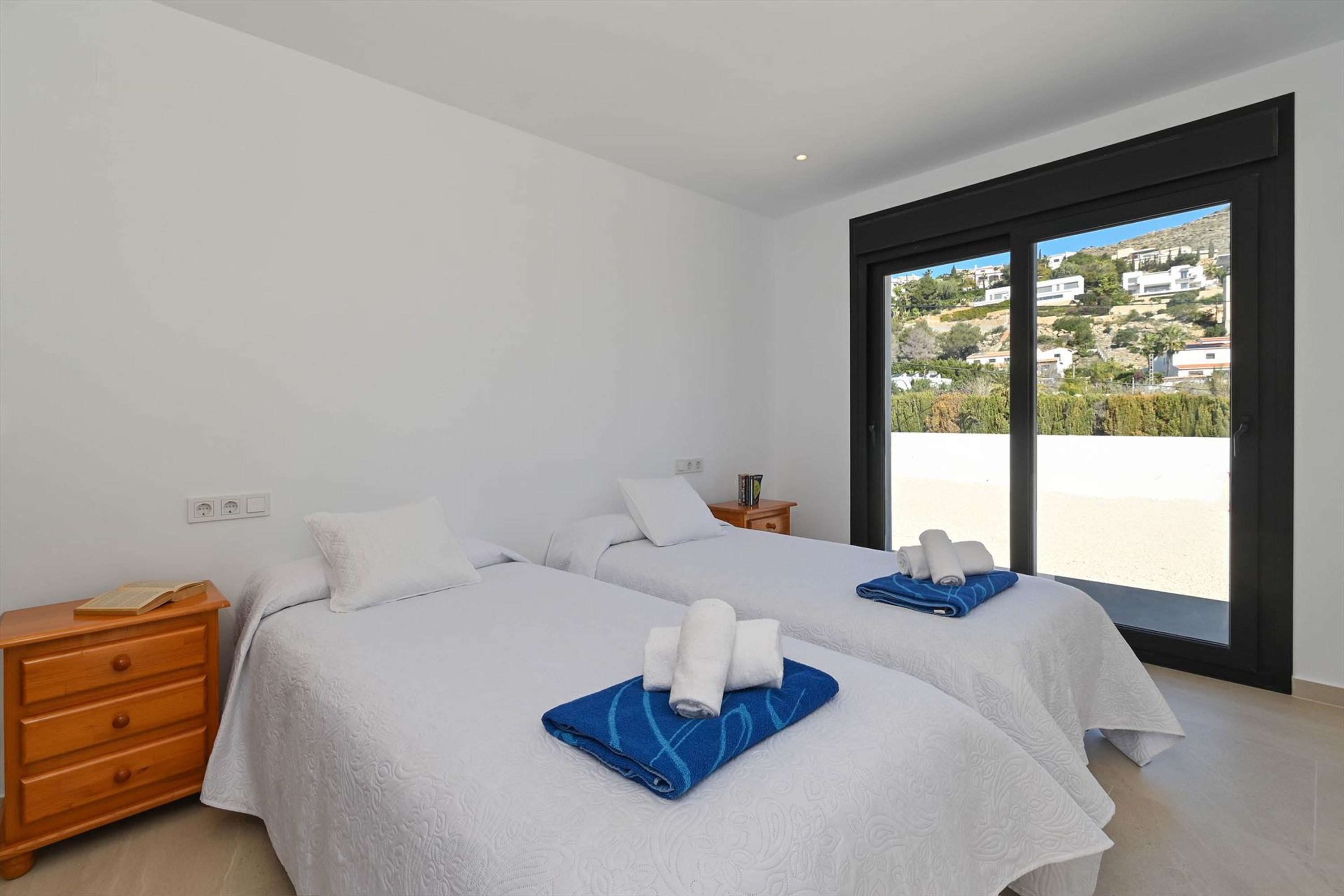 A hotel room with two beds, a wooden nightstand, and a large window offering a view of a hillside.