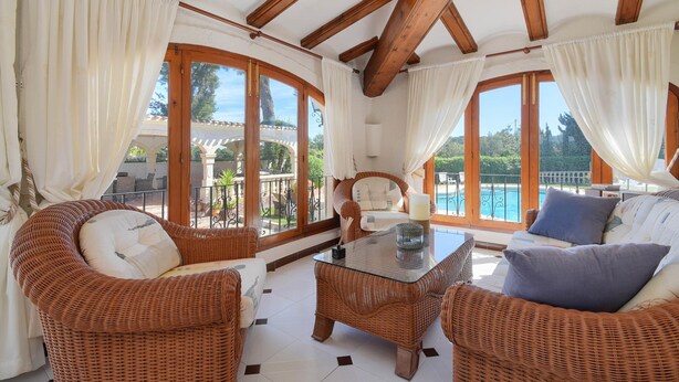 Ein sonnendurchflutetes Zimmer mit Korbmöbeln, großen Fenstern und Blick auf einen Pool.