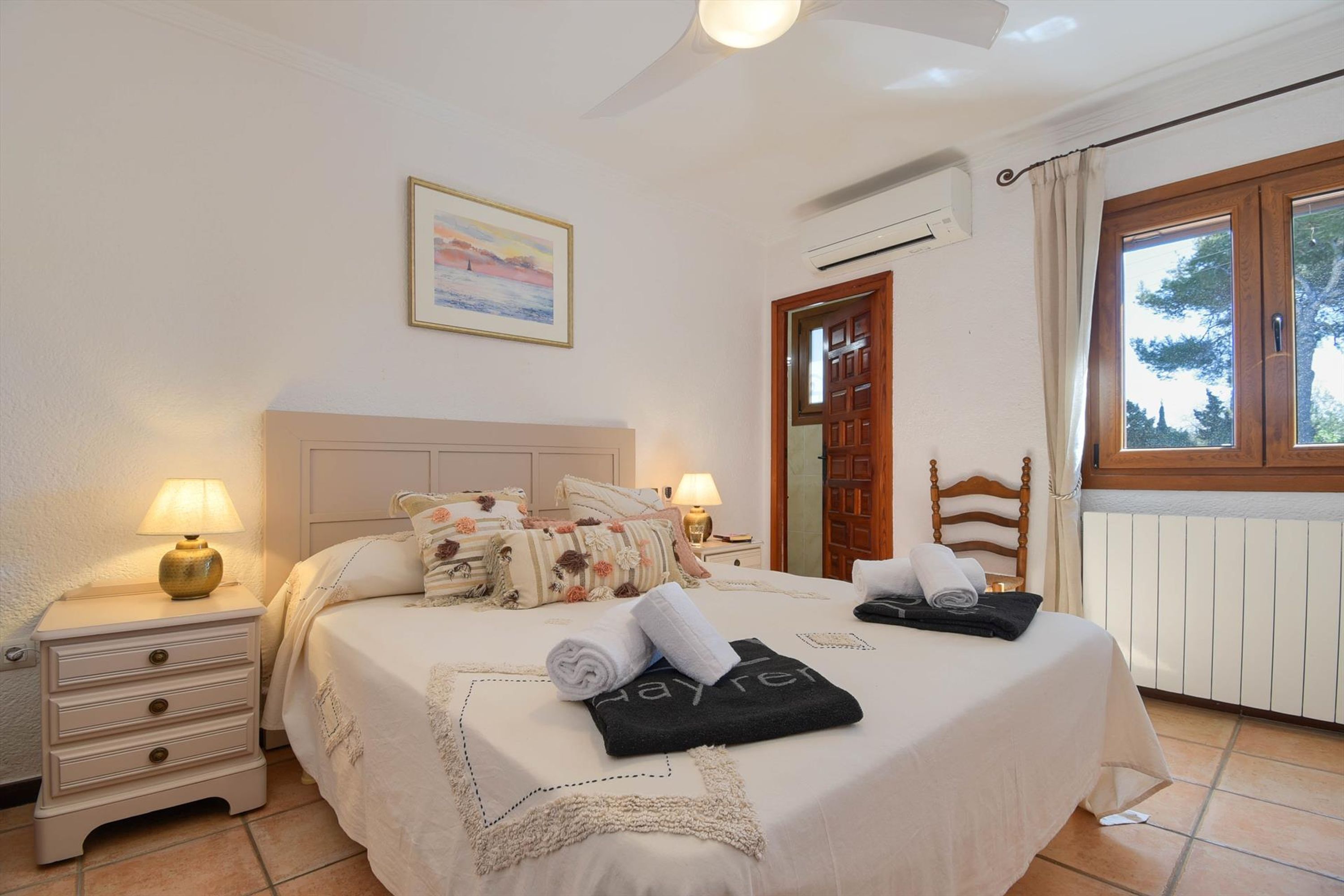 A neatly made bed with a white bedspread and decorative pillows, a bedside table with a lamp, and a window with curtains.
