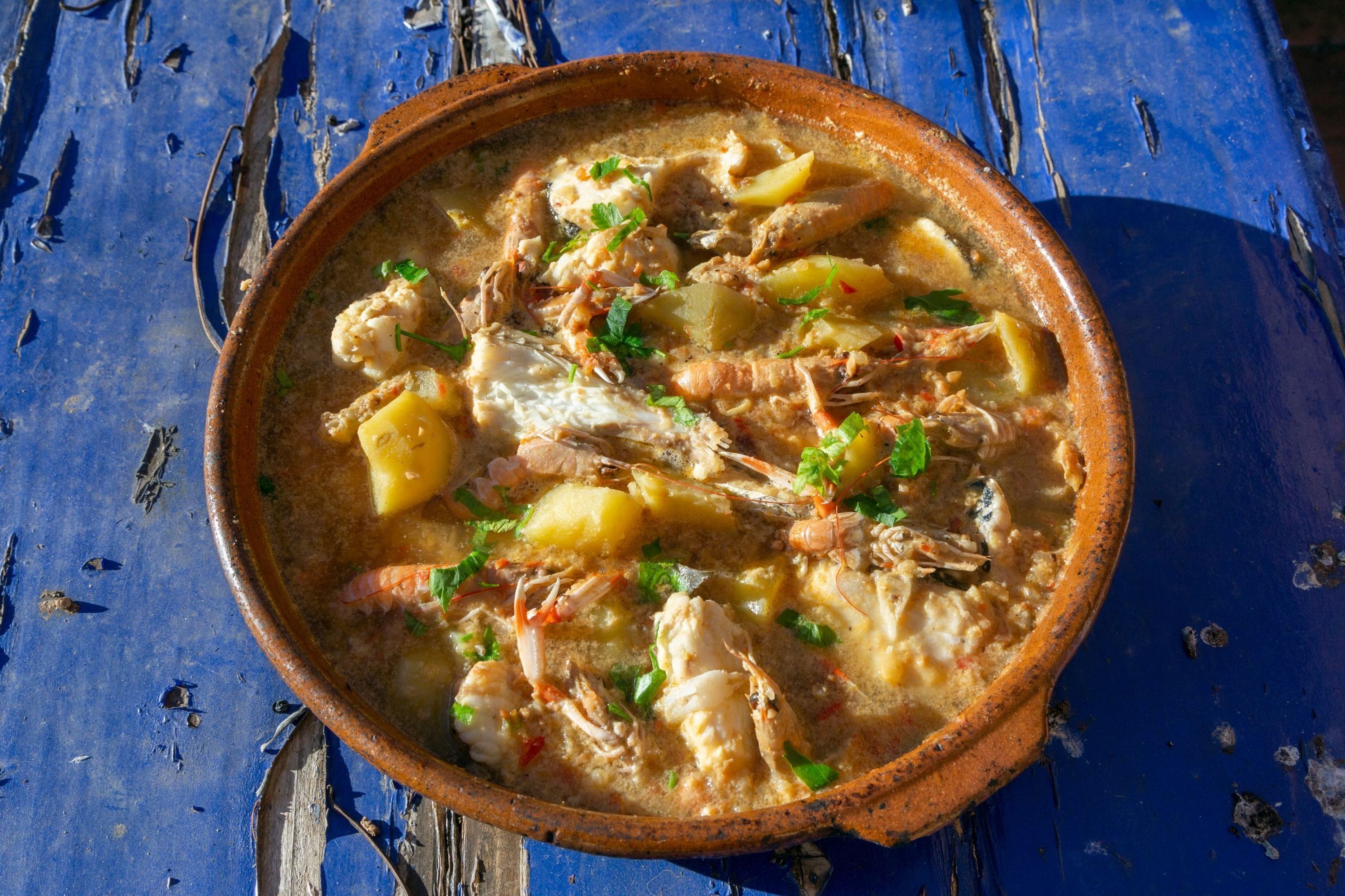 Un pot en terre cuite rustique, rempli d’une soupe de fruits de mer et garni d’herbes aromatiques, posé sur une surface en bois bleue.