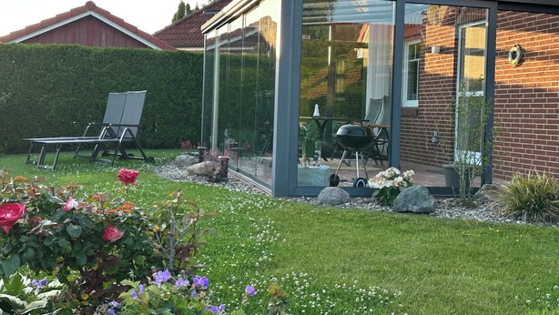 Eine moderne, ganzheitig verglaste Terrasse mit Grill, Sitzgelegenheit und gepflegtem Garten.
