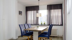 Ruokailutilassa on puinen pöytä ja sinisellä kankaalla verhoillut tuolit, riippuvalaisin sekä näkymä rakennuksiin suurten, verhoilla varustettujen ikkunoiden kautta.
