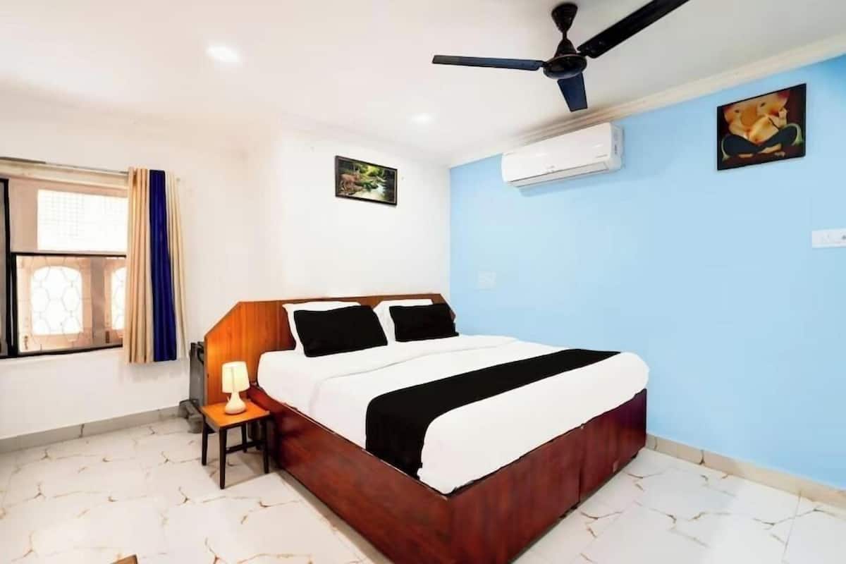 Un dormitorio con cama, ventilador de techo, equipo de aire acondicionado, ventana con cortinas y una mesita de noche con lámpara.
