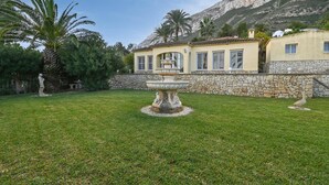 Un jardin bien entretenu, agrémenté d’une fontaine centrale, d’un mur en pierre et d’un bâtiment doté de grandes fenêtres et d’un toit en tuiles.