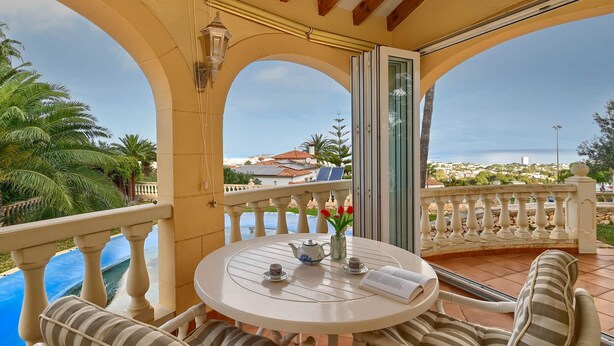 Une table ronde avec des coussins à rayures, une théière, des tasses et un livre, le tout placé sur un balcon donnant sur une piscine et offrant une vue sur la côte.