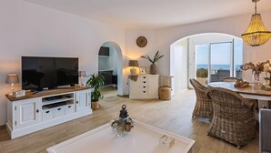 Een moderne woonkamer met een flatscreen televisie, witte kasten, een eethoek met rieten stoelen en uitzicht op zee.