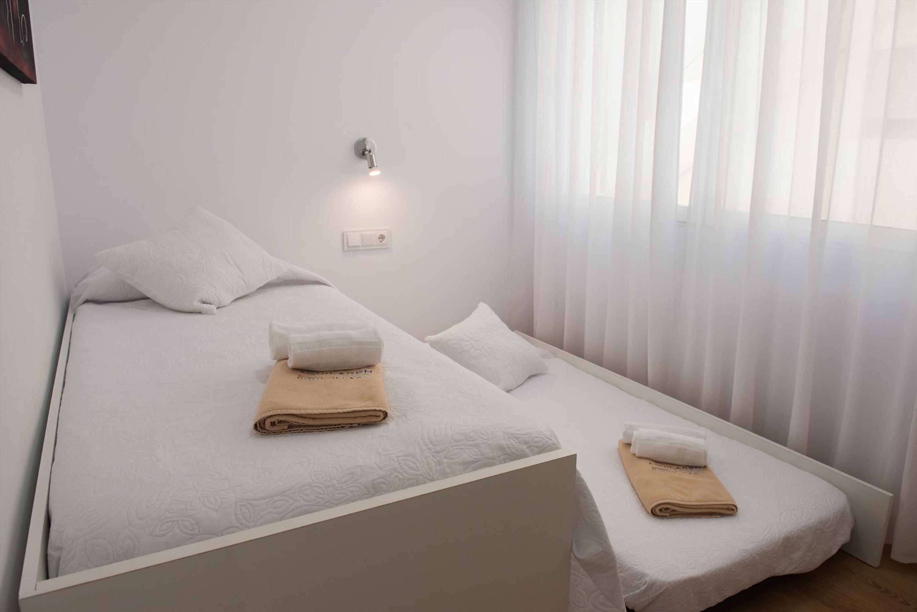 A small, neatly made twin bed with white bedding and two rolled towels, one beige and one white, in a bright room with a wall-mounted light and sheer curtains.