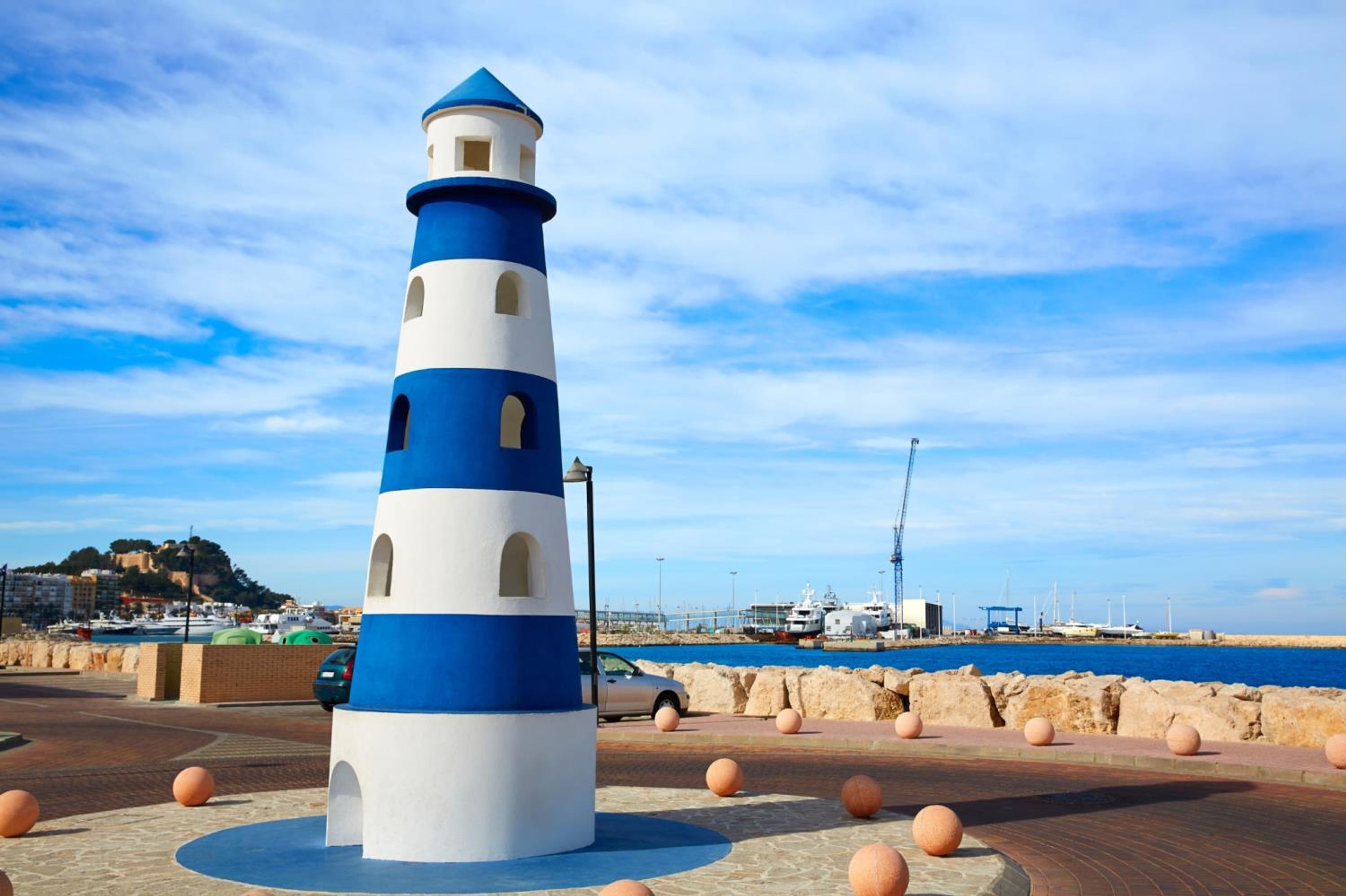 A lighthouse with a blue and white color scheme, located near a waterfront with boats and buildings in the background.