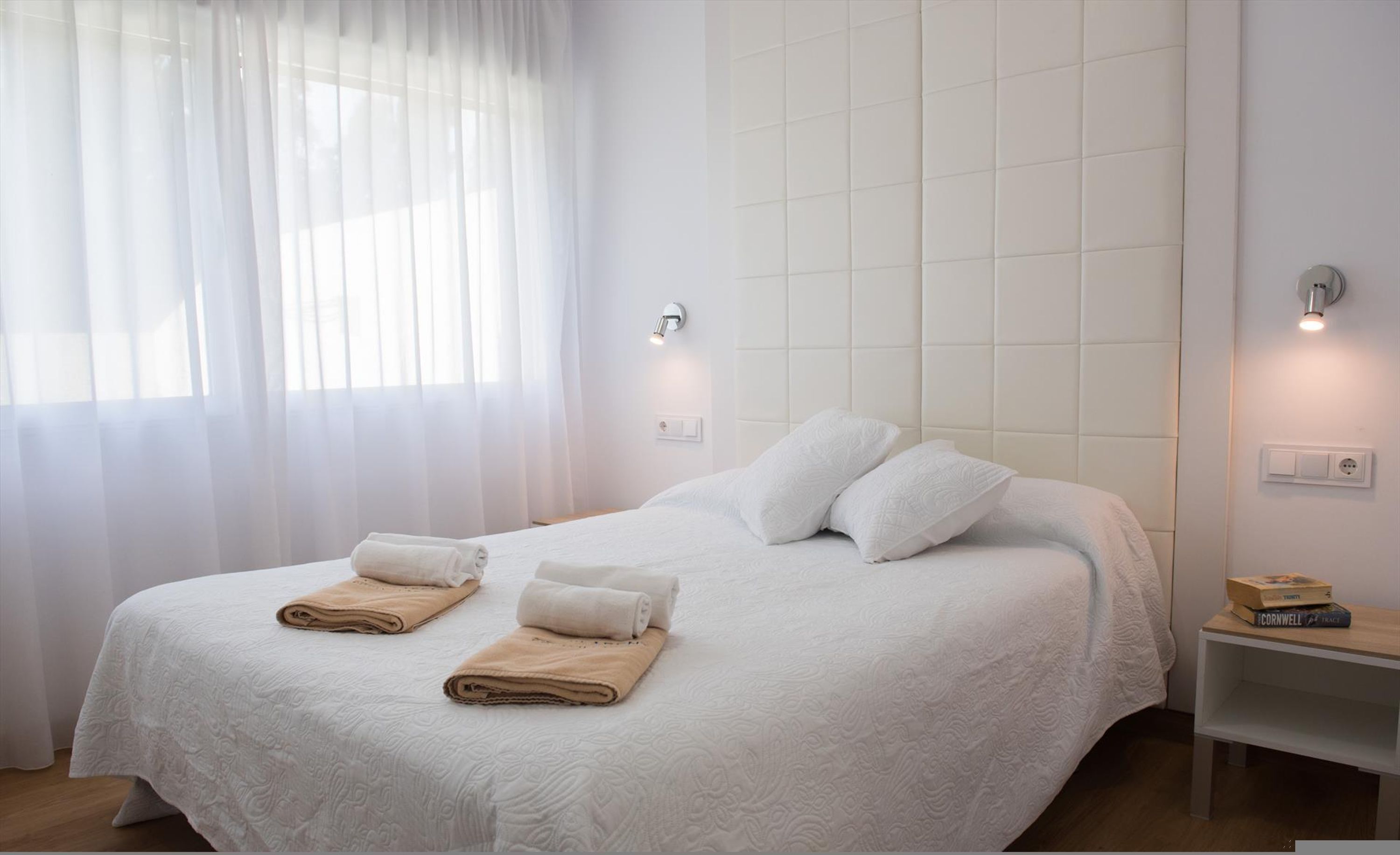 Un lit bien fait, avec du linge de lit blanc et deux serviettes beiges, une table de chevet avec un livre et un luminaire fixé au mur.