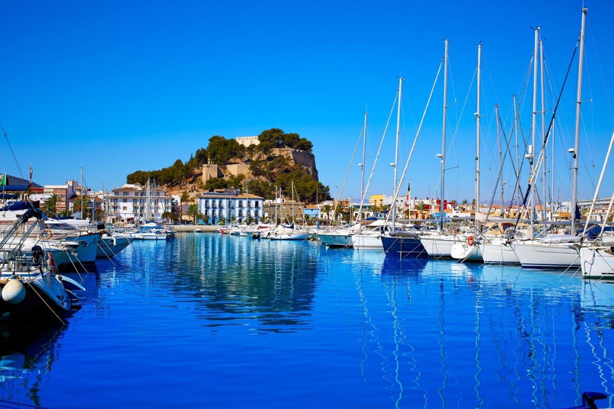Ein Yachthafen mit vertäuten Booten, ein markanter Hügel mit Gebäuden und strahlend blauem Himmel.