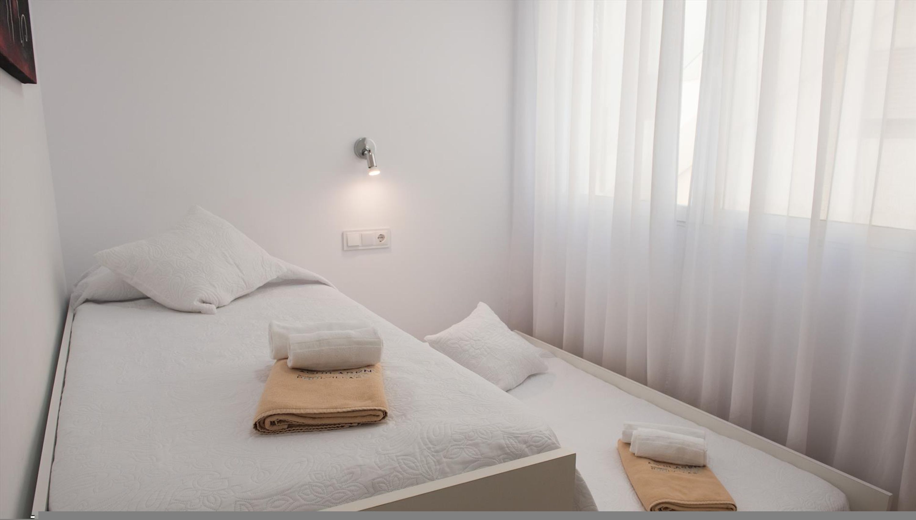 A single bed with white linens and two towels, a wall-mounted light, and sheer curtains.