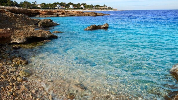 A coastal scene with clear blue waters, rocky shore, and buildings in the background. - Marina A3 4 pax in Balc n del Mar (Denia)