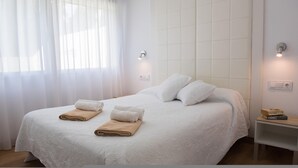 Una cama bien hecha con sábanas blancas y dos toallas color beige, una mesita de noche con un libro, y una lámpara fijada a la pared.