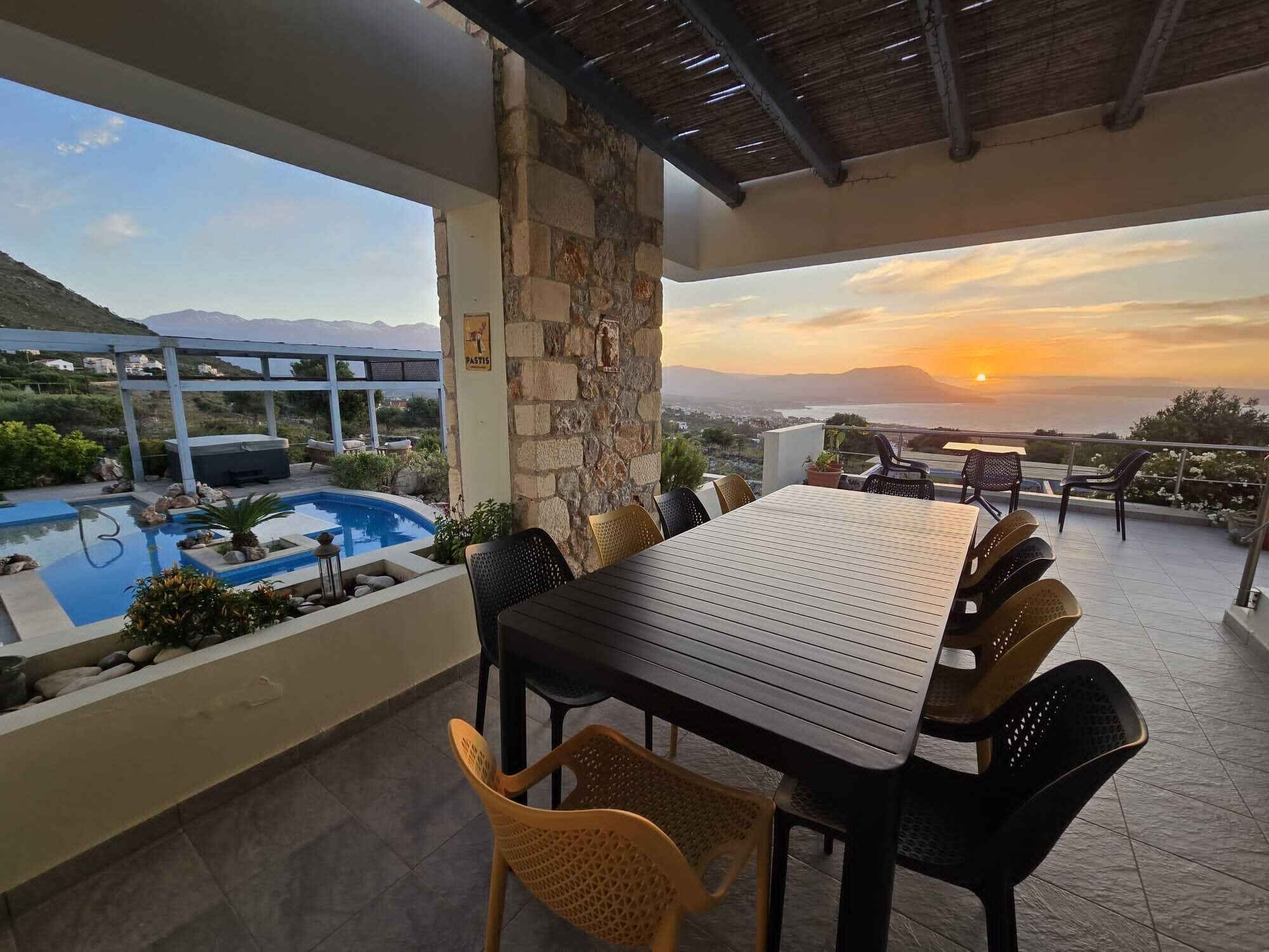 A terrace with a pool, dining table, and chairs, overlooking a scenic view of mountains and sunset.