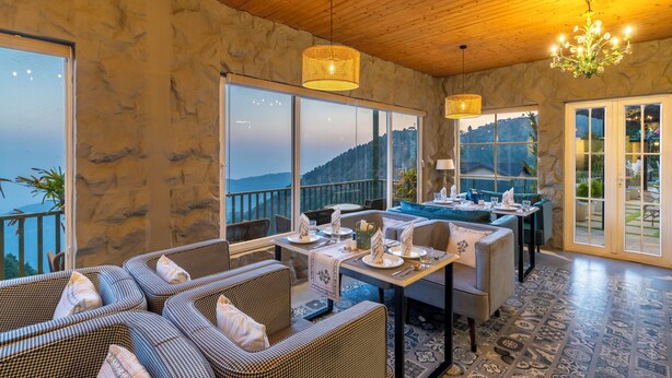 A dining area with wicker furniture, a chandelier, and large windows offering a view of the mountains. - The English Country House (Mussoorie)