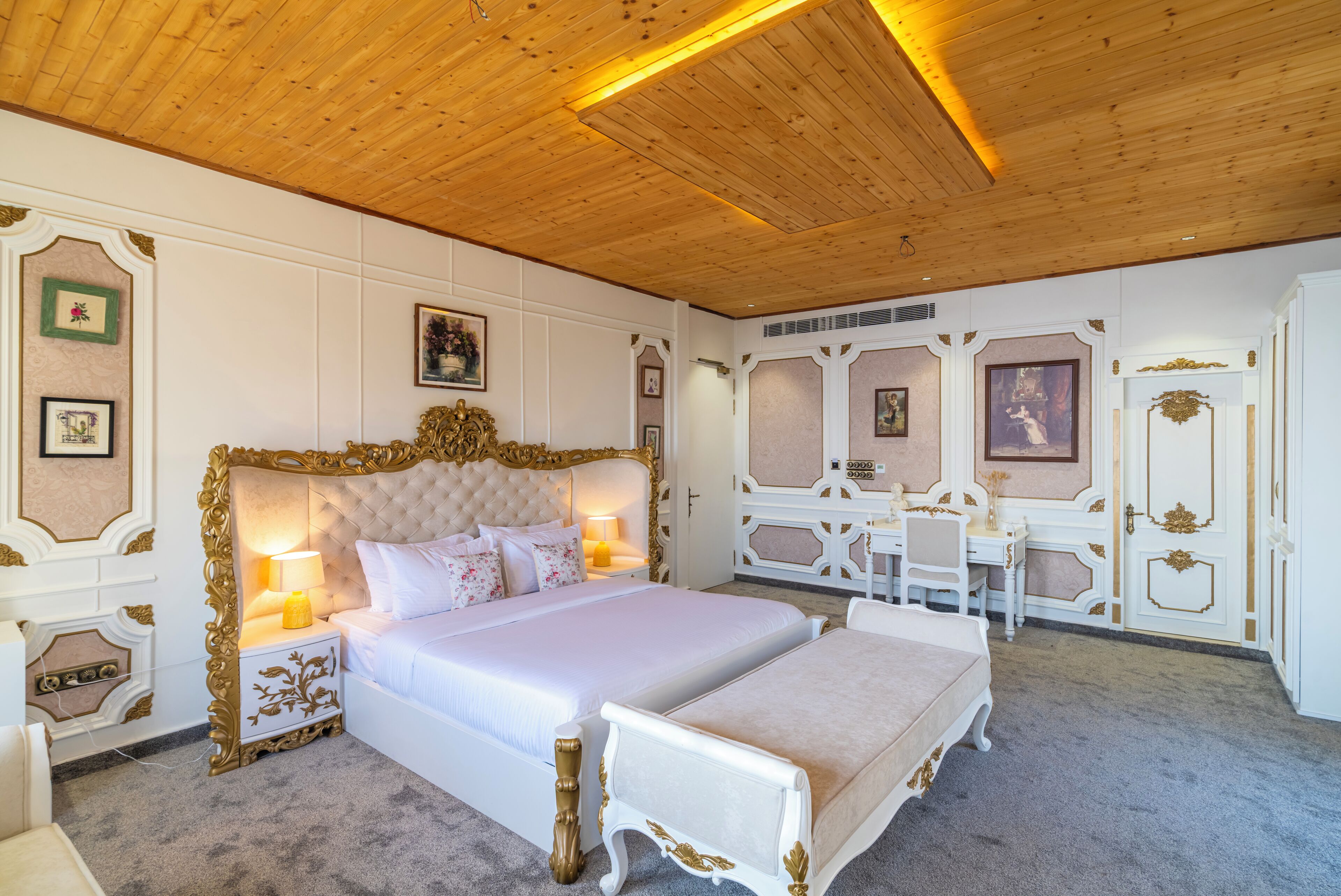 A large bed with a tufted headboard, bedside tables with lamps, and a small table with chairs in a room with wooden ceiling and white walls with decorative moldings.