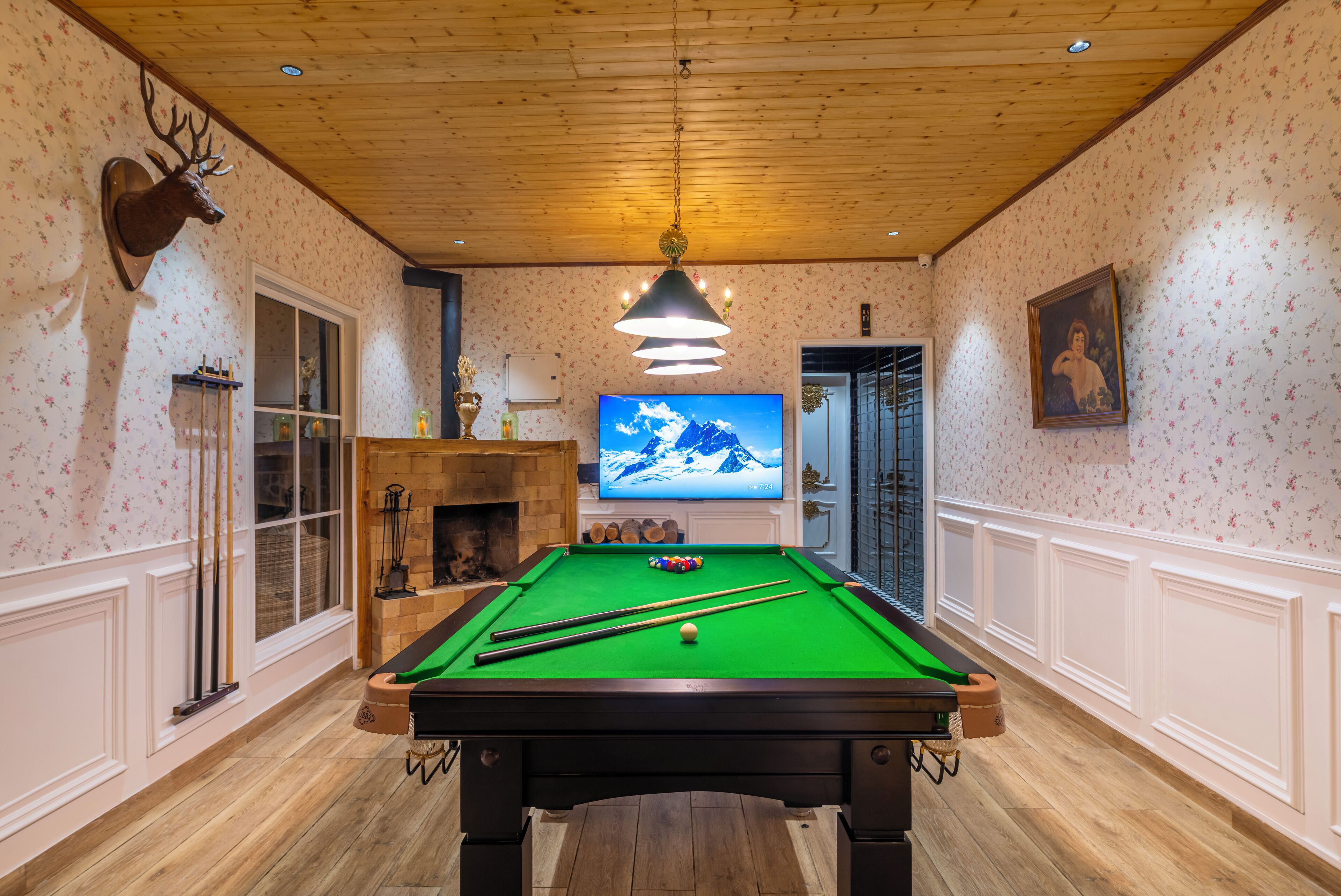 A pool table in a room with wooden flooring, a mounted deer head, a television, and a fireplace.