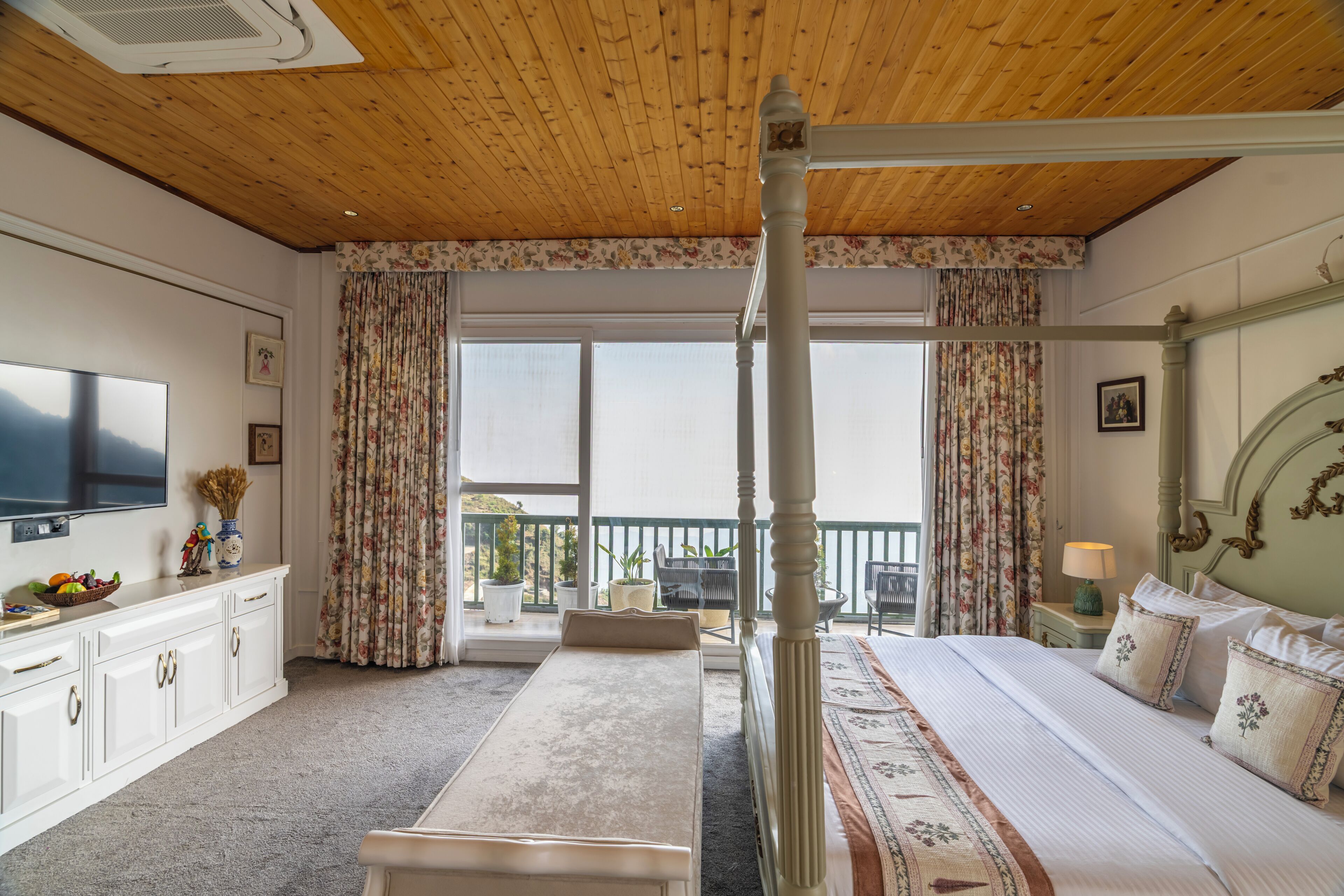 A bedroom with a four-poster bed, a television, a desk with a vase of flowers, and a balcony with curtains.