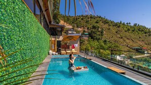 A swimming pool with a glass barrier, a wooden deck, and a view of a hillside. - The English Country House (Mussoorie)