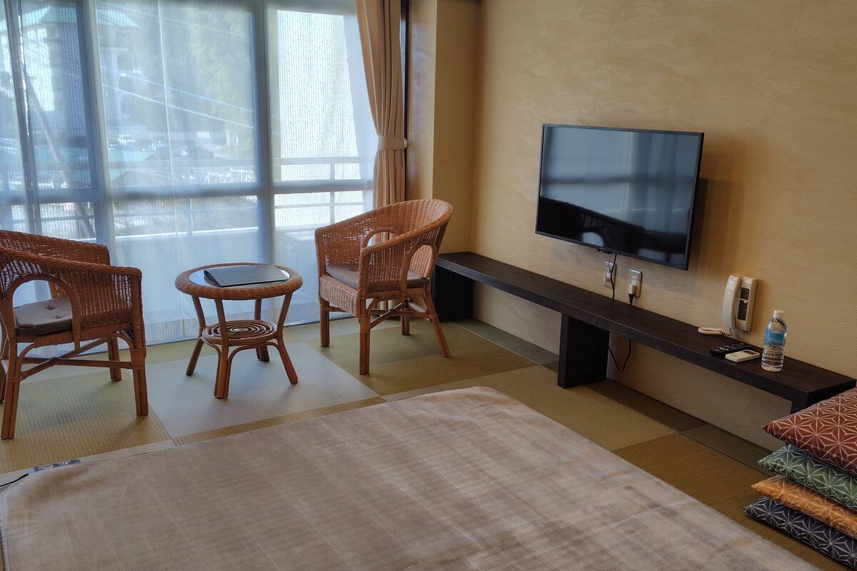 A hotel room with a television, wicker furniture, a large window, and a flat-screen TV mounted on the wall.