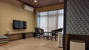 Habitación de hotel con televisión de pantalla plana, dos sillones de mimbre, una mesita, un refrigerador pequeño y una cama con almohadones estampados.