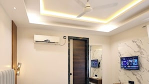 A modern hotel room with a large bed, ceiling fan, air conditioning unit, TV, marble wall, and a small shelf. - Hotel RGS Crystal (Kanpur)