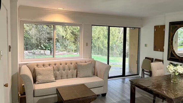 Un salon avec un canapé beige, une table basse en bois et une table à manger avec des chaises. De grandes fenêtres et une porte-fenêtre coulissante donnent sur un jardin.