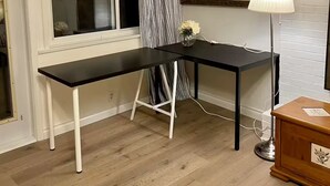 Une pièce avec un bureau noir, un lampadaire, une fenêtre avec des rideaux et un tableau accroché au mur.