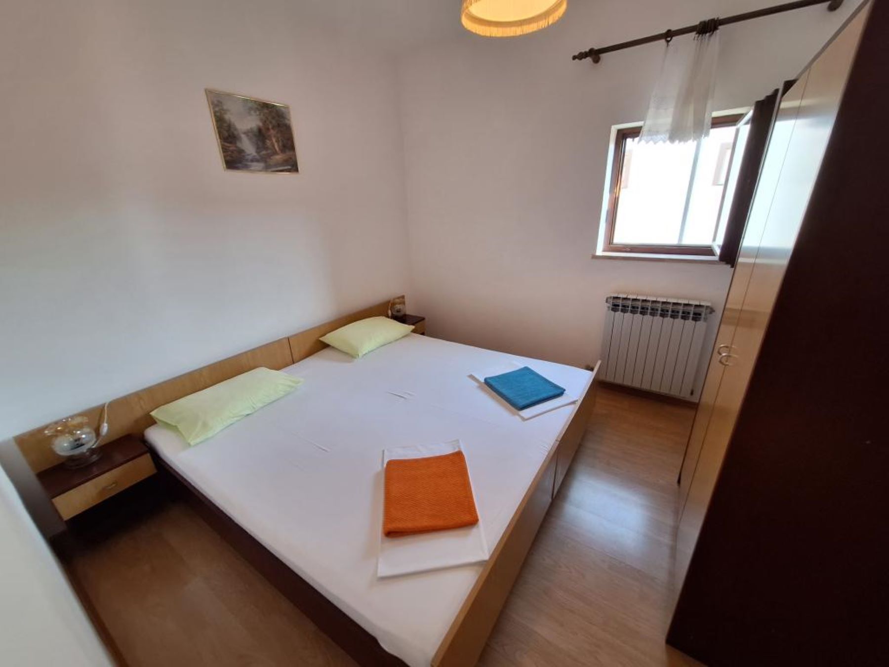 A single bed with a wooden headboard, a small bedside table, a window with curtains, and a radiator.