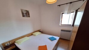 A single bed with a wooden headboard, a small bedside table, a window with curtains, and a radiator. - Apartman Štefica with AC and balcony for 2 people (Punat)